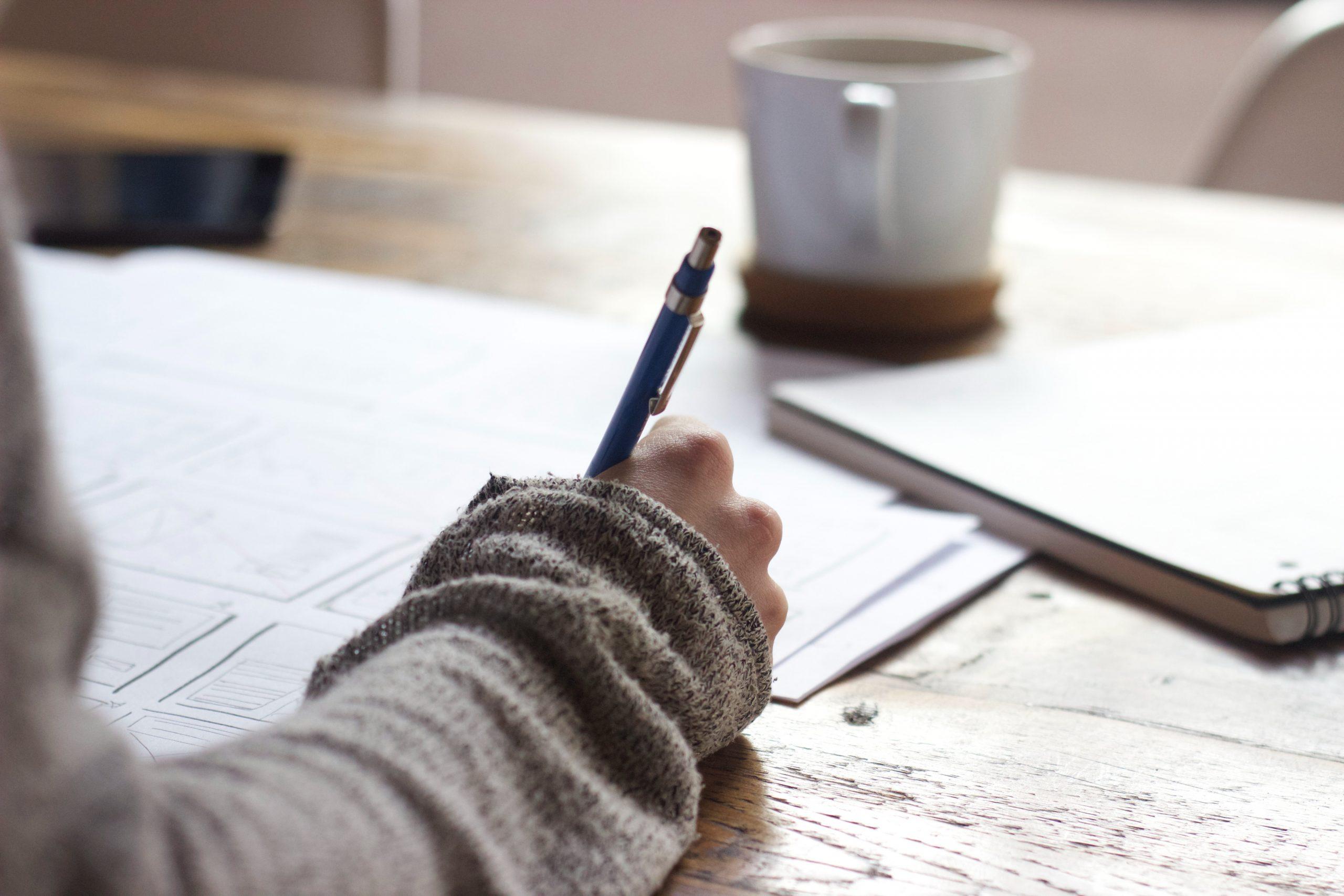 Person writing at a desk with a cup of coffee in the background