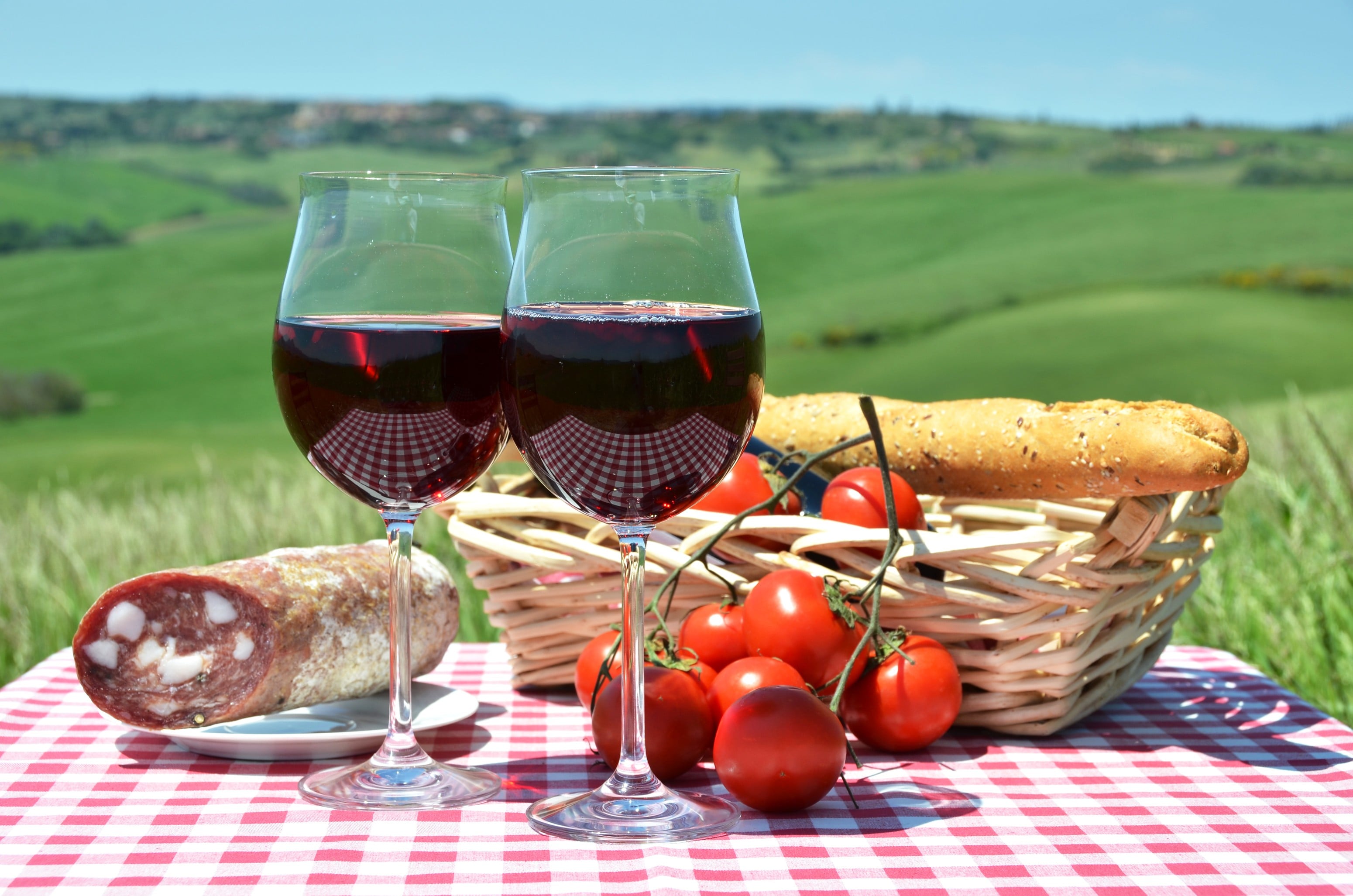 Picture of a picnic with two wine glasses filled with red wine, salami, tomatoes, and loaf of bread.